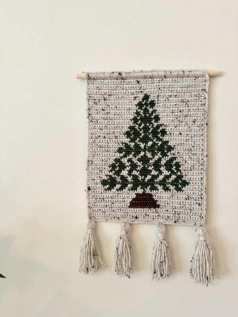 May include: A handmade, rectangular wall hanging featuring a crocheted Christmas tree design. The tree is dark green, set against a speckled gray background. The piece is finished with four cream-colored tassels and a wooden dowel for hanging.
