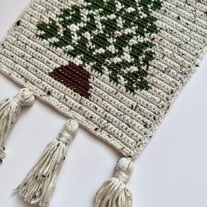 May include: Crocheted table runner featuring a green tree design on a speckled cream background. The tree has a brown trunk. The runner has three cream-colored tassels hanging from the bottom edge. The handmade runner is perfect for a rustic or farmhouse style.