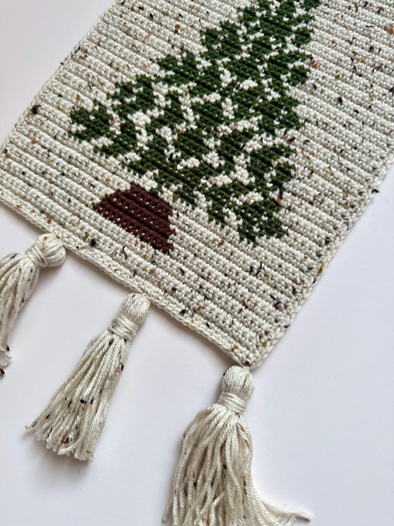 May include: Crocheted table runner featuring a green tree design on a speckled cream background. The tree has a brown trunk. The runner has three cream-colored tassels hanging from the bottom edge. The handmade runner is perfect for a rustic or farmhouse style.