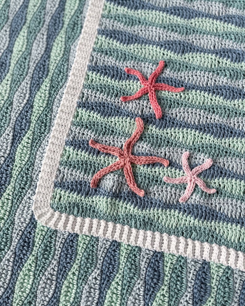 K&ouml;nnte beinhalten: Eine geh&auml;kelte Babydecke mit einem wellenf&ouml;rmigen Muster in Blaut&ouml;nen und Gr&uuml;n. Die Decke hat einen wei&szlig;en Rand und ist mit drei rosa geh&auml;kelten Seesternen verziert.