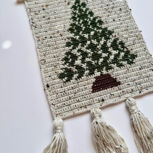 May include: A handmade, rectangular wall hanging featuring a crocheted Christmas tree design. The tree is green with a brown trunk, set against a speckled, off-white background. Three tassels hang from the bottom, and a wooden dowel is at the top.