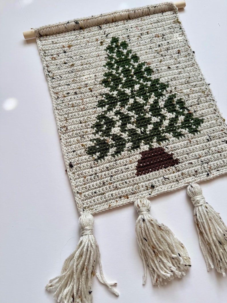 May include: A handmade, rectangular wall hanging featuring a crocheted Christmas tree design. The tree is green with a brown trunk, set against a speckled, off-white background. Three tassels hang from the bottom, and a wooden dowel is at the top.