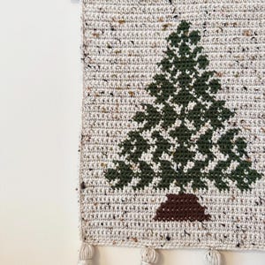 May include: A handmade crochet wall hanging featuring a green Christmas tree design on a speckled beige background. The tree has a brown trunk. The wall hanging is finished with tassels and a wooden dowel for hanging.