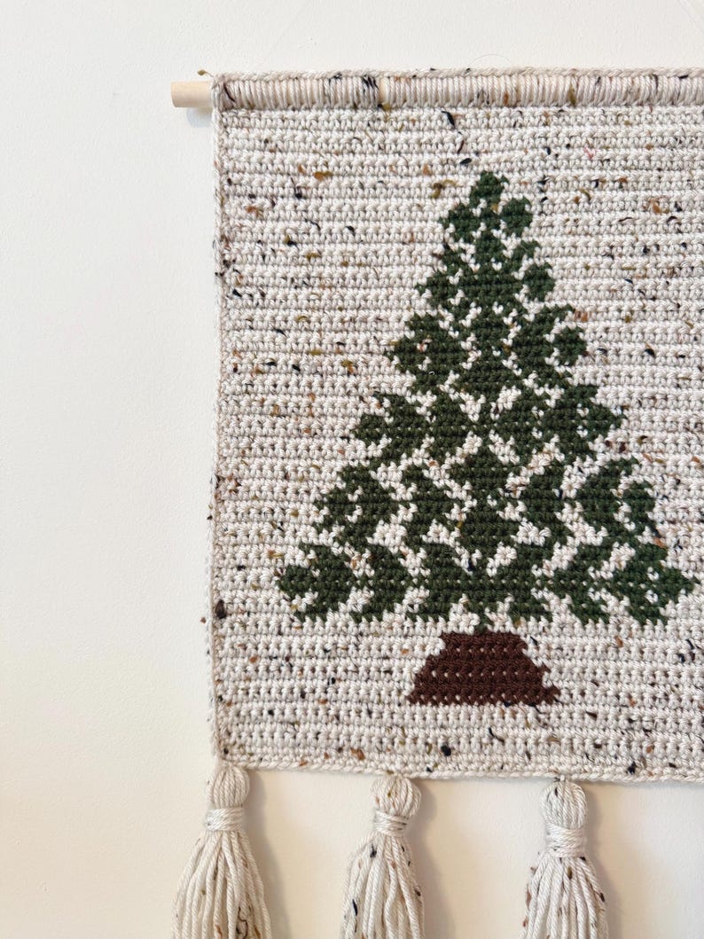 May include: A handmade crochet wall hanging featuring a green Christmas tree design on a speckled beige background. The tree has a brown trunk. The wall hanging is finished with tassels and a wooden dowel for hanging.