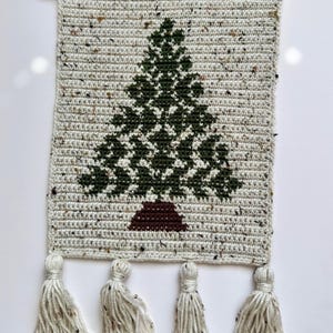 May include: Crocheted wall hanging featuring a green Christmas tree design on a speckled, off-white background. The tree has a brown base. The bottom edge has four tassels. A wooden dowel is at the top for hanging.