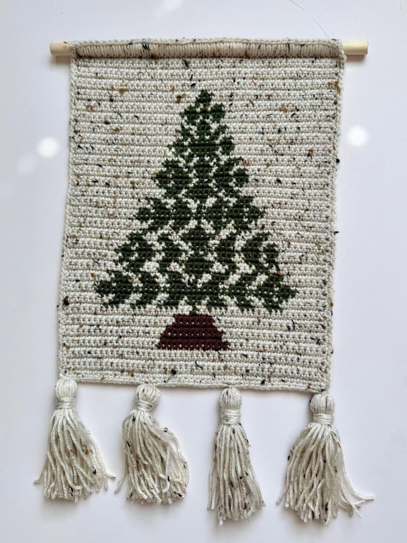 May include: Crocheted wall hanging featuring a green Christmas tree design on a speckled, off-white background. The tree has a brown base. The bottom edge has four tassels. A wooden dowel is at the top for hanging.