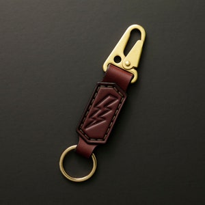 以下が含まれることがあります： ゴールドの金属製の留め具と稲妻のデザインが施された、茶色の革製のキーホルダー。キーホルダーには、キーを取り付けるためのゴールドの金属製のリングが付いています。