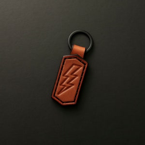 以下が含まれることがあります： 黒い金属製のリングと稲妻のデザインが施された茶色の革製のキーホルダー
