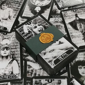 Puede incluir: Una baraja de cartas del tarot con imágenes en blanco y negro, con imágenes de películas vintage y bordes de estilo art déco. La baraja está sujeta con una banda verde y sellada con un sello de cera dorado. Las cartas están esparcidas.
