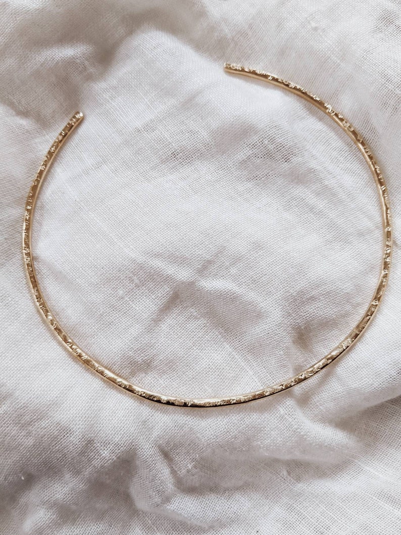 May include: A gold choker necklace with a textured finish. The necklace is lying on a white fabric background.