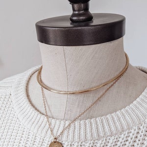 May include: A gold chain necklace with a round pendant on a white sweater. The necklace is layered over a gold choker.