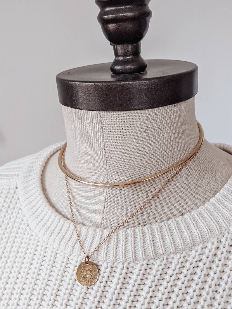 May include: A gold chain necklace with a round pendant on a white sweater. The necklace is layered over a gold choker.