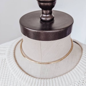 May include: A gold choker necklace on a white mannequin. The necklace is a simple, thin band of metal.