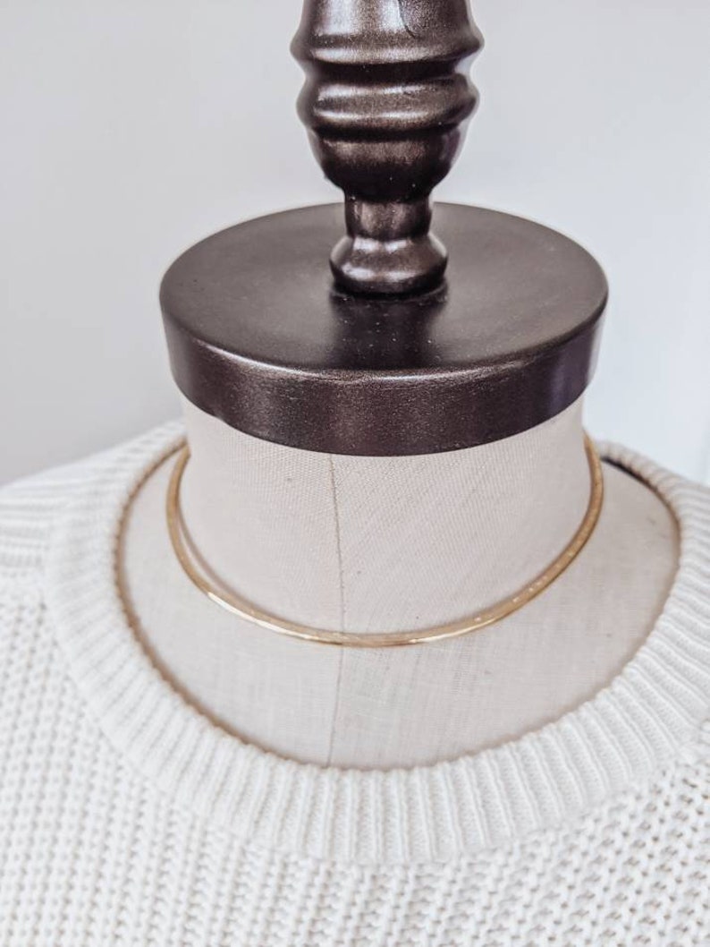 May include: A gold choker necklace on a white mannequin. The necklace is a simple, thin band of metal.
