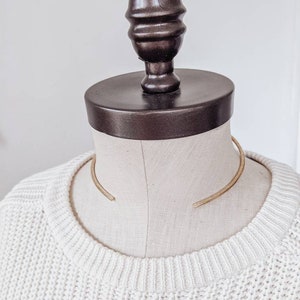 May include: A gold metal choker necklace on a white mannequin. The necklace is a simple, curved design.
