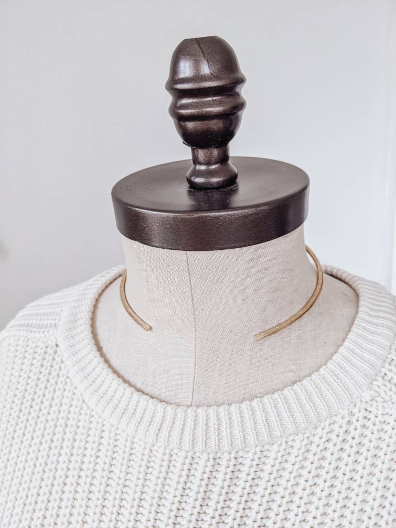 May include: A gold metal choker necklace on a white mannequin. The necklace is a simple, curved design.
