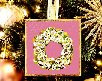 Op de afbeelding: Een vierkante kerstboomversiering met een roze achtergrond en een geschilderd kransontwerp in tinten groen, bruin en wit. De versiering heeft een goudkleurige rand en een wit lintlusje aan de bovenkant om het ornament op te hangen.