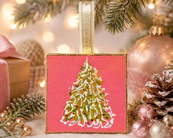 Puede incluir: Un adorno colgante cuadrado con un fondo de lienzo rosa vibrante y un árbol de Navidad pintado en verde, decorado con líneas blancas en espiral y detalles dorados, acabado con un borde dorado y un lazo de cinta de tela color crema.