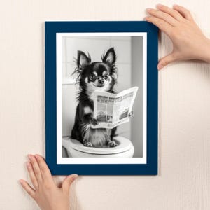 May include: A black and white photo of a small dog sitting on a toilet and reading a newspaper. The dog is wearing a collar and has a happy expression on its face. The photo is framed in a dark blue frame.