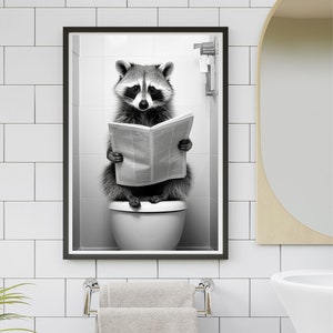 以下が含まれることがあります： トイレに座って新聞を読んでいるアライグマの白黒写真。写真は黒の額縁に入っていて、白いタイル張りの壁にかけられています。
