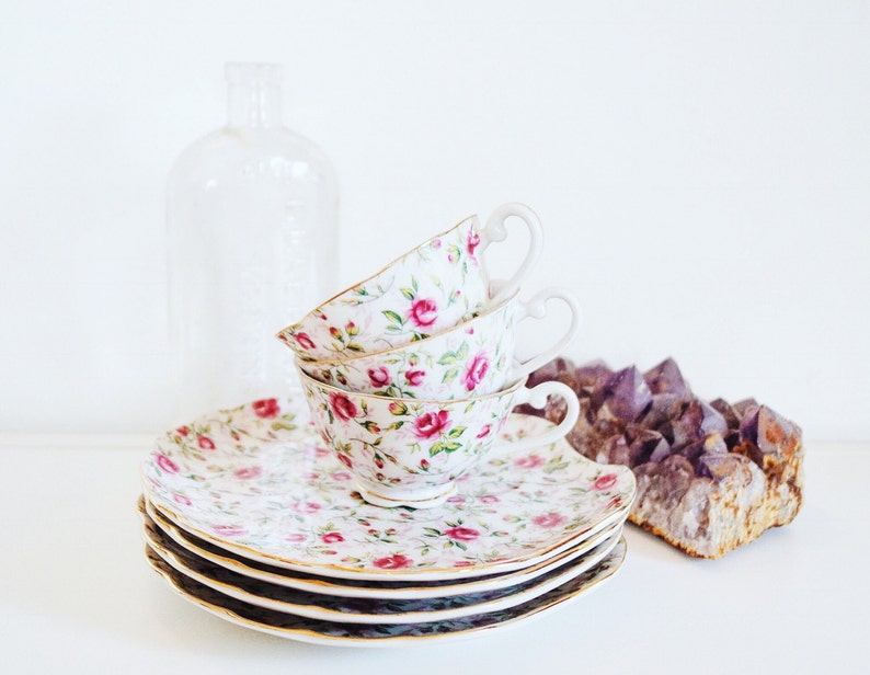May include: A set of four floral teacups and saucers with a pink and white floral pattern. The cups are stacked on top of each other and the saucers are stacked underneath. The cups and saucers are on a white surface.