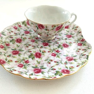 May include: A white porcelain teacup and saucer set with a floral pattern of pink roses and green leaves. The saucer has a scalloped edge and a gold trim.