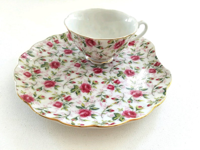 May include: A white porcelain teacup and saucer set with a floral pattern of pink roses and green leaves. The saucer has a scalloped edge and a gold trim.