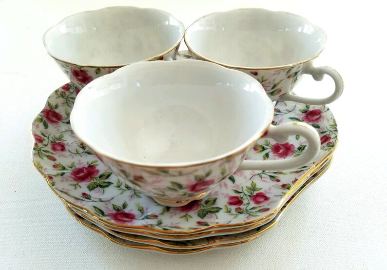May include: Three white teacups with floral patterns and gold trim, stacked on top of each other. The cups are on a floral patterned saucer.