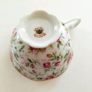 May include: A white teacup with a floral pattern of pink roses and green leaves. The bottom of the cup has a gold crown and the text "Carlton China, Hand Painted, Made in Portugal, July 14, 1964".