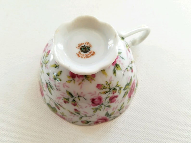 May include: A white teacup with a floral pattern of pink roses and green leaves. The bottom of the cup has a gold crown and the text "Carlton China, Hand Painted, Made in Portugal, July 14, 1964".
