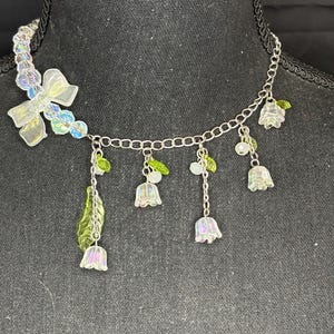 May include: A delicate necklace featuring a silver chain with iridescent flower and leaf charms. The necklace includes a bow and clear beads. The flowers and leaves have green and clear accents, creating a whimsical and elegant design.