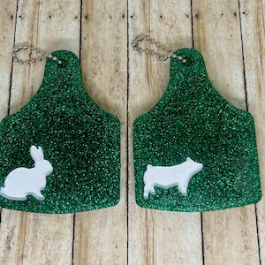 May include: Two green glittery tags with silver chain attachments. One tag features a white rabbit, the other a white pig. The tags are likely for livestock or animal identification.
