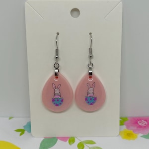 May include: A pair of pink teardrop-shaped earrings with a bunny rabbit inside a blue and purple patterned egg. The earrings are on a white card with a hole at the top.