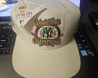 Puede incluir: Una gorra de béisbol blanca New Era con un parche de la Serie Mundial 1996 Campeones con el logotipo de los New York Yankees. El parche está bordado con detalles coloridos y un bate de béisbol.