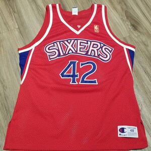 May include: Red basketball jersey with blue and white trim. The jersey has "SIXERS" written across the chest in white with blue outlines. The number "42" is printed in blue on the front of the jersey. The jersey is made by Champion and has a size tag on the bottom right side.