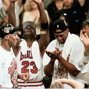 May include: Four men wearing white shirts and hats are celebrating. The man in the center is wearing a red and white basketball jersey with the number 23 on it. The jersey says "Bulls" on the front.