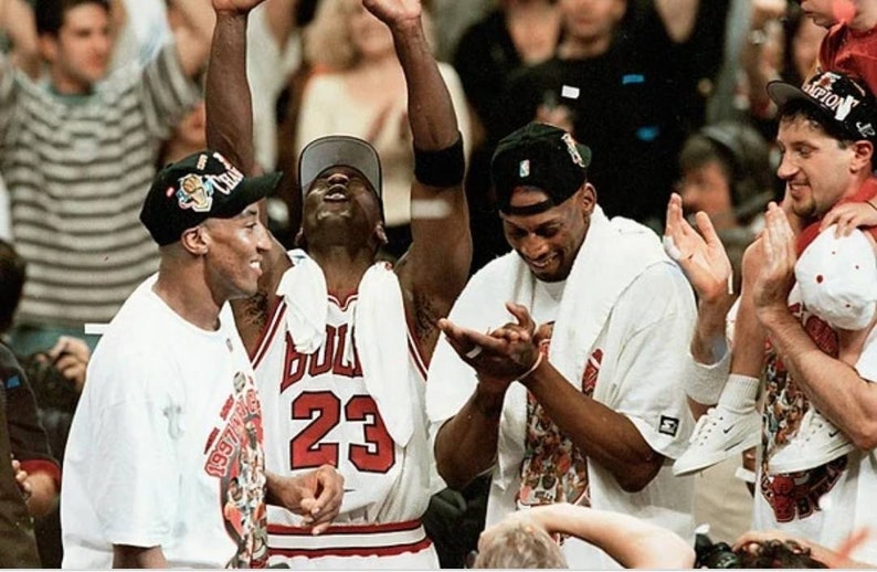 May include: Four men wearing white shirts and hats are celebrating. The man in the center is wearing a red and white basketball jersey with the number 23 on it. The jersey says "Bulls" on the front.