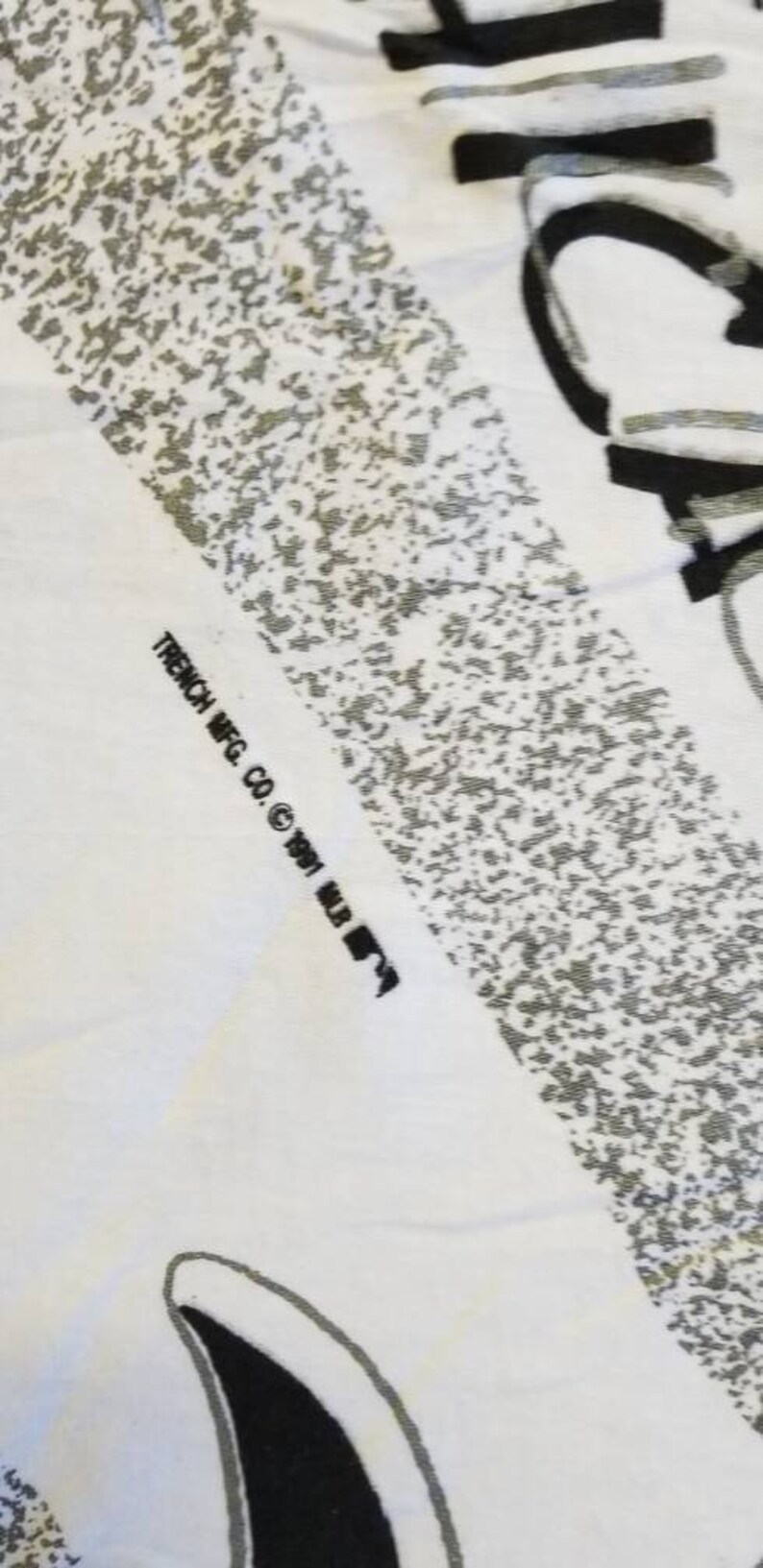 Puede incluir: Una camiseta blanca con un estampado gr&aacute;fico en blanco y negro. El texto "TRENCH MFG. CO. &copy; 1991 MLB" est&aacute; impreso en la parte inferior de la camiseta.