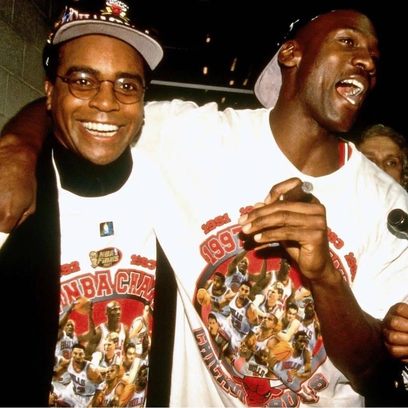 May include: Two men wearing white t-shirts with a red and white graphic of the 1997 NBA Champions Chicago Bulls. The man on the left is smiling and the man on the right is holding a cigar and smiling.