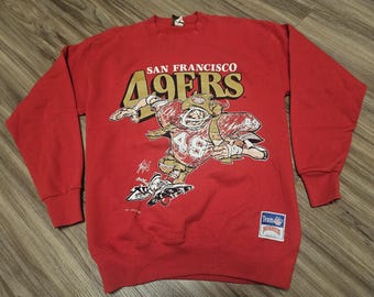 Puede incluir: Sudadera roja de manga larga con una ilustración de dibujos animados de un jugador de fútbol americano que lleva una camiseta de los San Francisco 49ers. El texto "San Francisco 49ers" está impreso en la parte delantera de la sudadera.