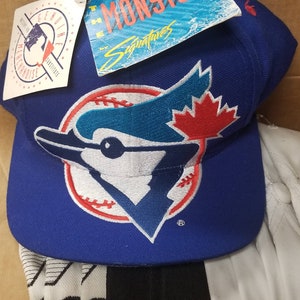 May include: A royal blue baseball cap featuring the Toronto Blue Jays logo, a stylized blue jay with a red maple leaf. The cap has a flat brim and a white and black striped pattern on the underside. Tags are attached.