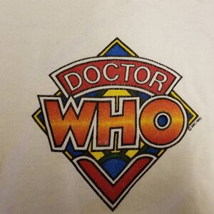 May include: White long-sleeve shirt with a colorful Doctor Who logo. The logo is a diamond shape with the words "Doctor Who" in a red, orange, yellow, and blue font. The text "c 1981-1983" is in the bottom right corner of the logo.
