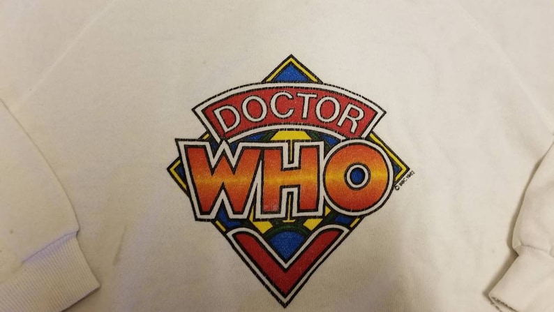 May include: White long-sleeve shirt with a colorful Doctor Who logo. The logo is a diamond shape with the words "Doctor Who" in a red, orange, yellow, and blue font. The text "c 1981-1983" is in the bottom right corner of the logo.