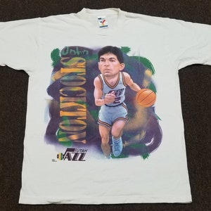 May include: White t-shirt with a cartoon illustration of John Stockton, a basketball player for the Utah Jazz. The shirt has the team logo and the player's name.