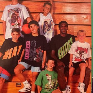 Op de afbeelding: Een groep jonge jongens die basketbaltruitjes dragen met verschillende NBA-spelers erop. De jongens zitten op een houten bank met een basketbal voor zich. Een van de jongens draagt een Celtics-trui met de tekst "CELTICS" op de voorkant. Een andere jongen draagt een wit shirt met de tekst "JORDAN" op de voorkant. De jongens lachen allemaal en kijken naar de camera.