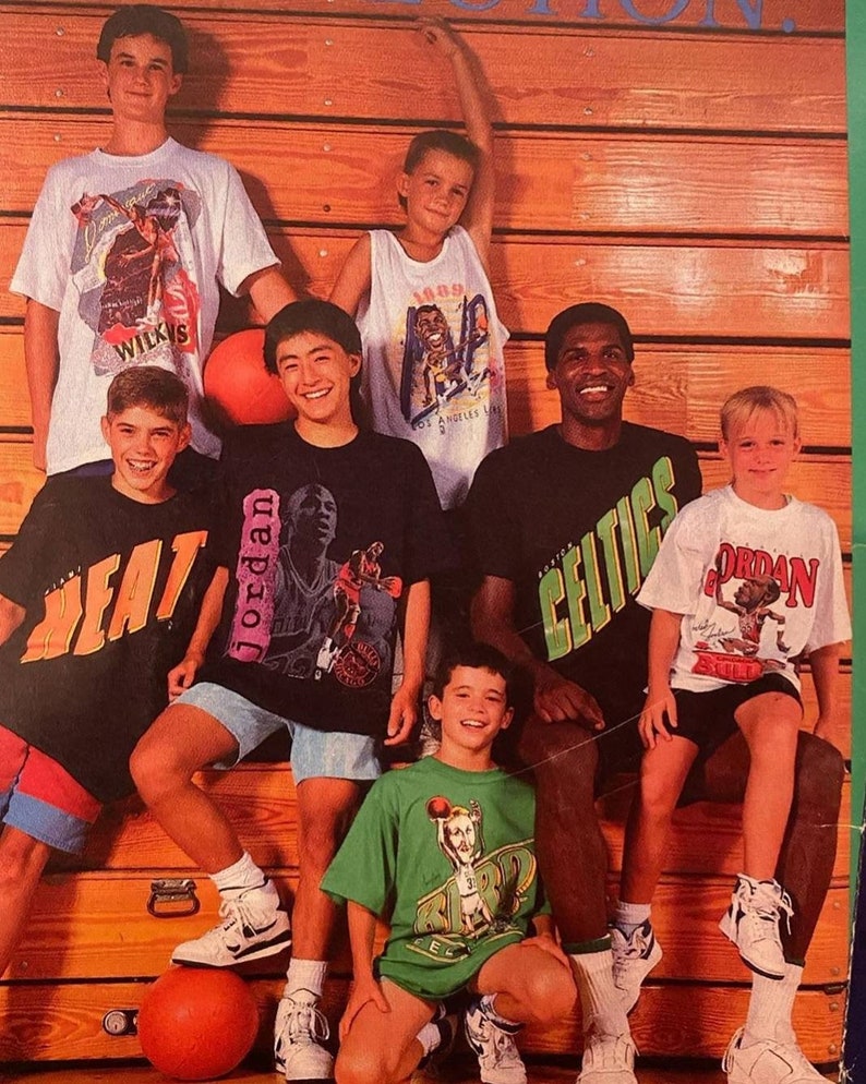 Op de afbeelding: Een groep jonge jongens die basketbaltruitjes dragen met verschillende NBA-spelers erop. De jongens zitten op een houten bank met een basketbal voor zich. Een van de jongens draagt een Celtics-trui met de tekst "CELTICS" op de voorkant. Een andere jongen draagt een wit shirt met de tekst "JORDAN" op de voorkant. De jongens lachen allemaal en kijken naar de camera.