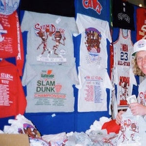 Op de afbeelding: Een verzameling vintage basketbal T-shirts met verschillende NBA-teamlogo's en spelers, waaronder de Orlando Magic, de Cleveland Cavaliers en de San Antonio Spurs. De shirts worden getoond op een blauwe muur met een witte achtergrond. Een persoon houdt een grijs T-shirt vast met een cartoon basketbalspeler afbeelding en de tekst "Slam Dunk Championship".