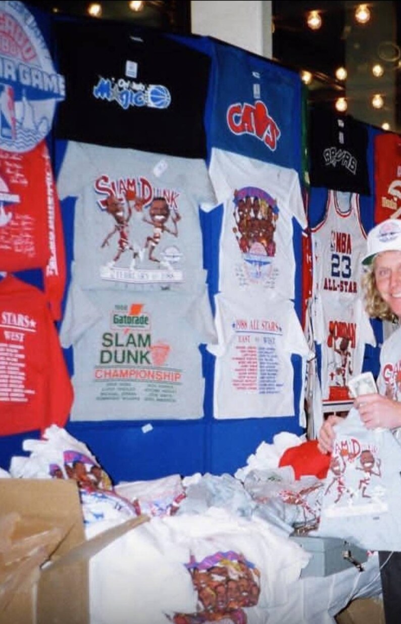Op de afbeelding: Een verzameling vintage basketbal T-shirts met verschillende NBA-teamlogo's en spelers, waaronder de Orlando Magic, de Cleveland Cavaliers en de San Antonio Spurs. De shirts worden getoond op een blauwe muur met een witte achtergrond. Een persoon houdt een grijs T-shirt vast met een cartoon basketbalspeler afbeelding en de tekst "Slam Dunk Championship".