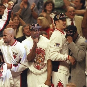 May include: A group of five basketball players wearing white shirts with a red and black logo and the text "Chicago Bulls" on the front. The players are celebrating a victory and are holding up their hands in the air. The players are wearing white and black hats with the text "Champions" on them.