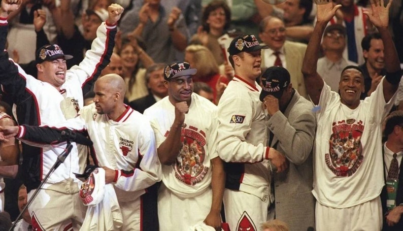 May include: A group of five basketball players wearing white shirts with a red and black logo and the text "Chicago Bulls" on the front. The players are celebrating a victory and are holding up their hands in the air. The players are wearing white and black hats with the text "Champions" on them.